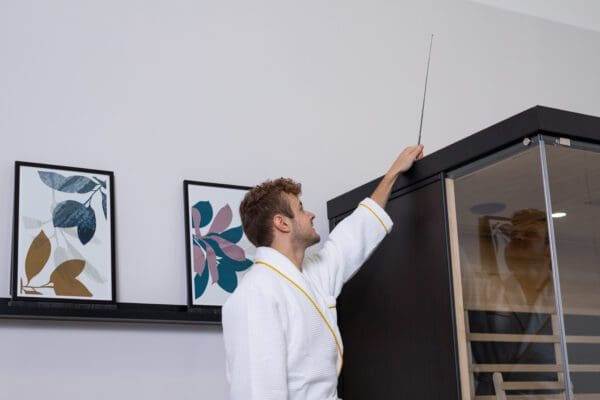 A man in a robe is adjusting the door of a Sirona 3-Person Hemlock Infrared Sauna with 8 Carbon Heaters.